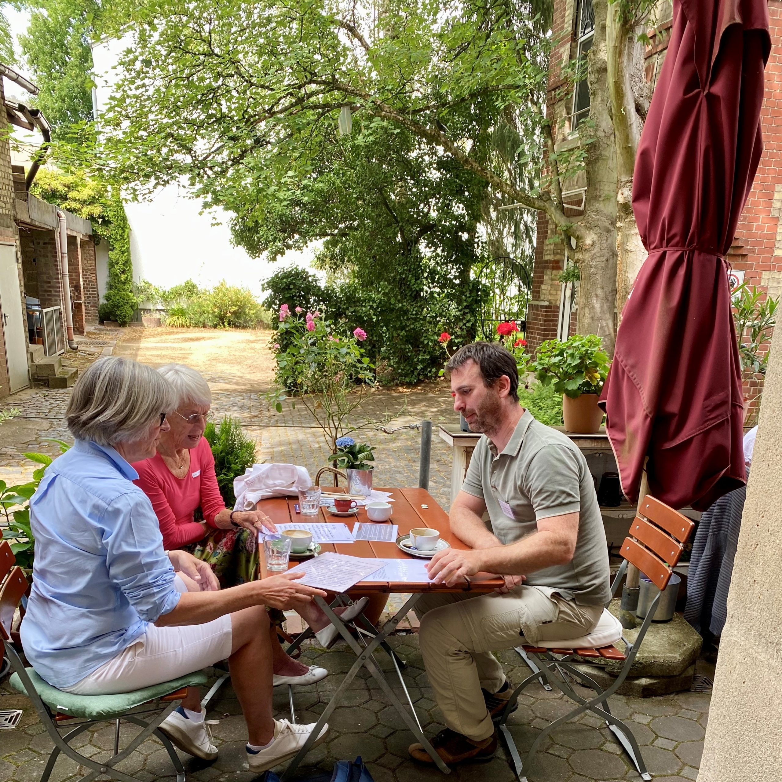 Ein wissenschaftlicher Mitarbeiter des „Gruß & Kuss“-Projekts diskutiert mit zwei interessierten Bürgerinnen über Liebesbriefe an einem Tisch auf der Terrasse des See You Cafés
